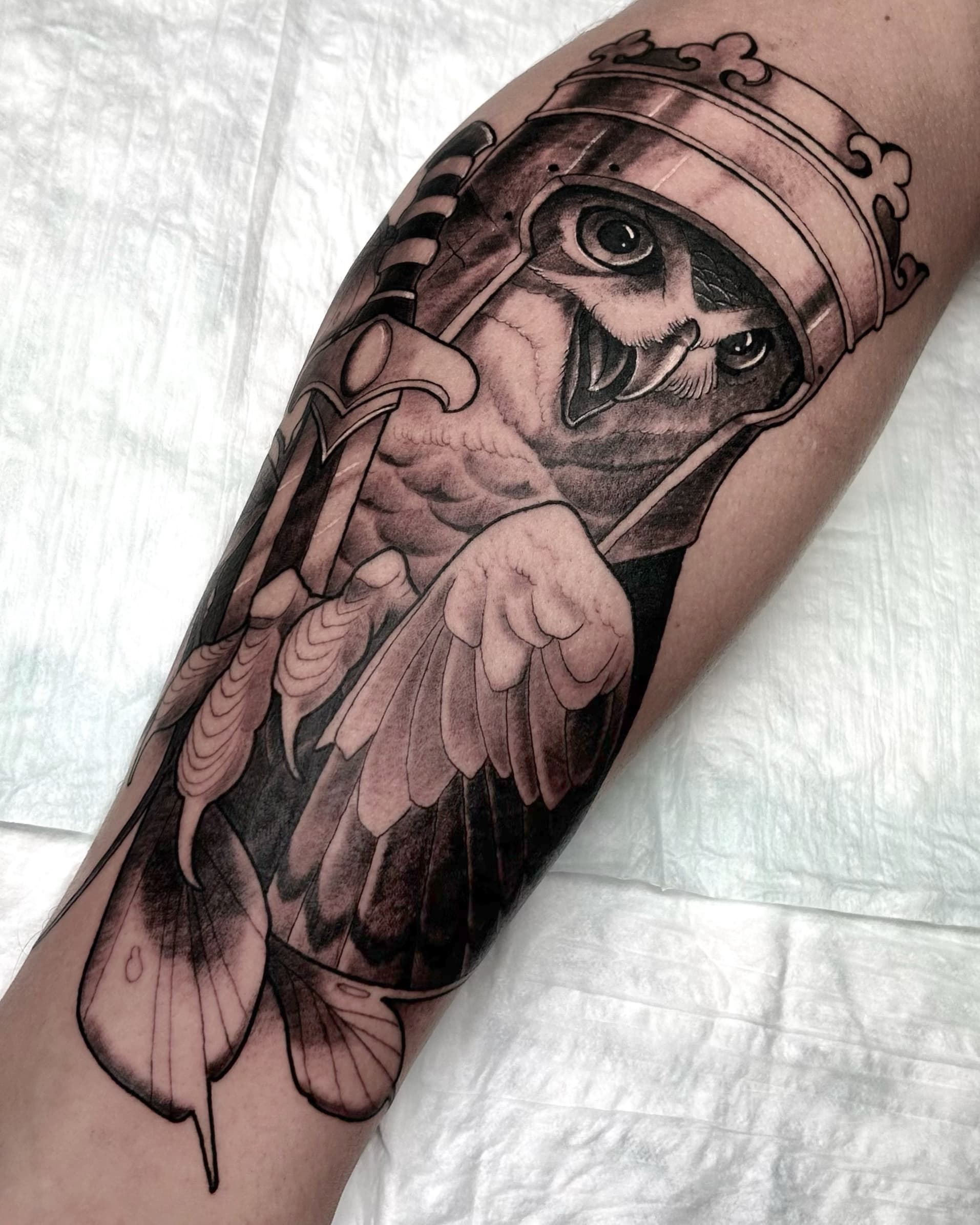Black and grey tattoo of an owl wearing a knight helmet and holding a sword — detailed illustrative design